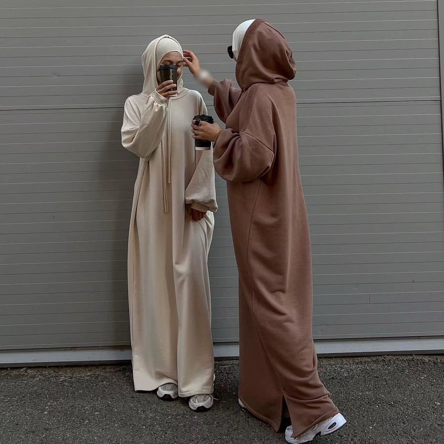 Zwei stilvoll gekleidete Frauen tragen moderne, minimalistische Abayas mit Kapuze in neutralen Tönen – eine in Cremeweiß, die andere in warmem Braun. Beide kombinieren ihre Outfits mit weißen Sneakers und tragen Sonnenbrillen. Sie genießen ihren Kaffee und eine der Frauen richtet sanft die Kopfbedeckung der anderen. Der schlichte, graue Hintergrund unterstreicht den modernen und eleganten Look.