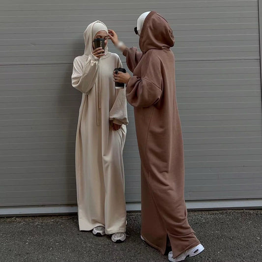 Zwei stilvoll gekleidete Frauen tragen moderne, minimalistische Abayas mit Kapuze in neutralen Tönen – eine in Cremeweiß, die andere in warmem Braun. Beide kombinieren ihre Outfits mit weißen Sneakers und tragen Sonnenbrillen. Sie genießen ihren Kaffee und eine der Frauen richtet sanft die Kopfbedeckung der anderen. Der schlichte, graue Hintergrund unterstreicht den modernen und eleganten Look.