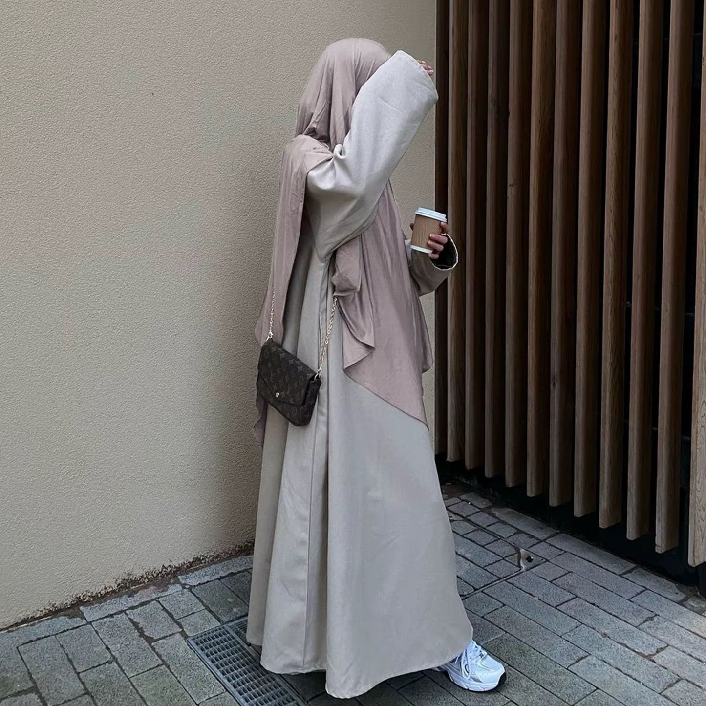 Eine stilvoll gekleidete Frau trägt eine fließende beige Abaya mit einem passenden Khimar in einem sanften Taupe-Ton. Sie hält einen Kaffeebecher in der Hand und trägt eine elegante, kleine Umhängetasche mit goldener Kette. Ihre weißen Sneaker verleihen dem Look eine moderne Note. Die dezente, neutrale Farbpalette des Outfits harmoniert mit dem minimalistischen, urbanen Hintergrund aus Holz und Beton.
