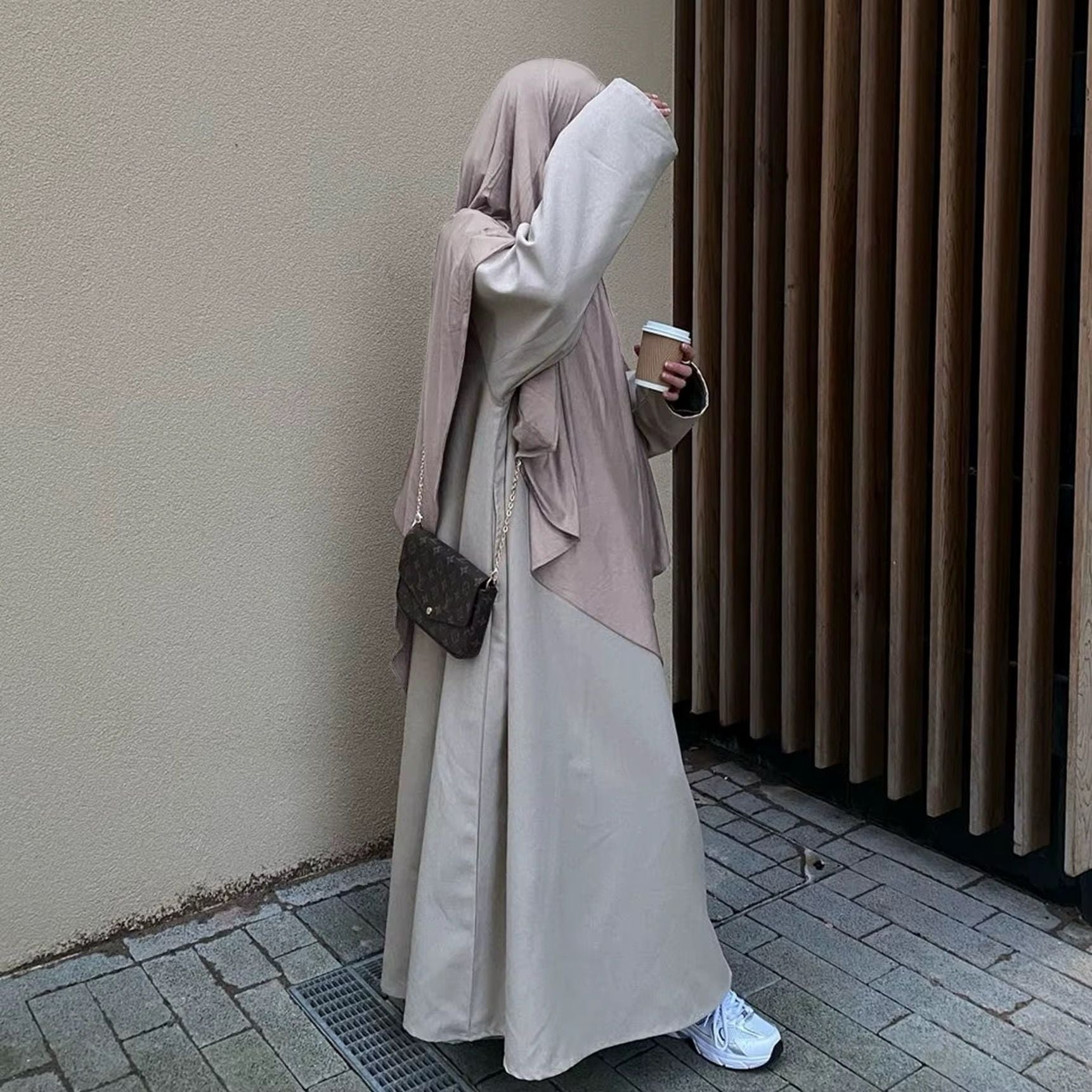 Eine stilvoll gekleidete Frau trägt eine fließende beige Abaya mit einem passenden Khimar in einem sanften Taupe-Ton. Sie hält einen Kaffeebecher in der Hand und trägt eine elegante, kleine Umhängetasche mit goldener Kette. Ihre weißen Sneaker verleihen dem Look eine moderne Note. Die dezente, neutrale Farbpalette des Outfits harmoniert mit dem minimalistischen, urbanen Hintergrund aus Holz und Beton.
